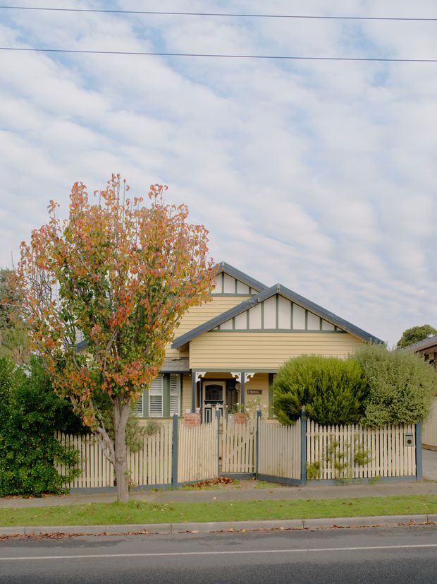 Street-facing view of a detached period-style house with pale weatherboard cladding, decorative gabled roof detailing, a small covered front porch, and a timber picket fence. Mature planting and an autumnal tree frame the facade, with overhead utility wires and a suburban street in the foreground.