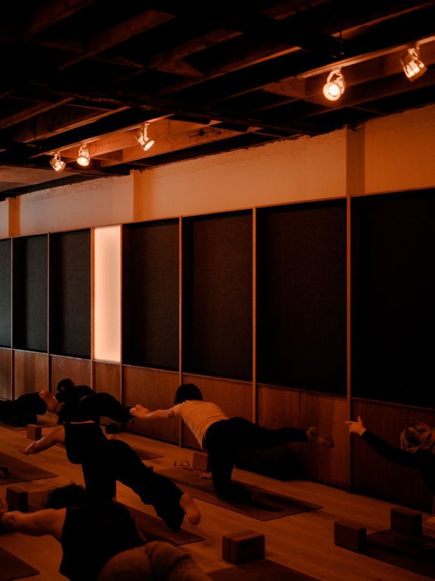 A dimly lit interior studio with a warm amber atmosphere, where several people are practicing yoga or floor-based exercise on individual mats. The room features dark timber floorboards, timber lower wall panelling, tall dark acoustic-looking wall panels, exposed ceiling beams, and track-mounted spotlights.