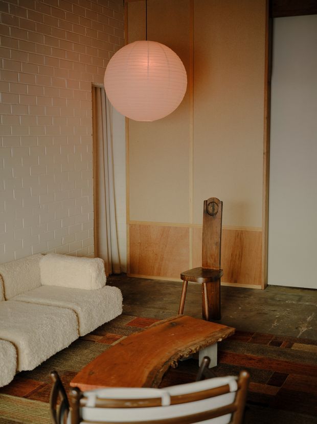 Warm, eclectic living room interior with a cream boucle sofa, sculptural timber coffee table, vintage wooden chair, large paper globe pendant, textured white brick wall, timber wall panelling, and dark patterned flooring. The room combines tactile upholstery and richly coloured natural wood with a relaxed mid-century-inspired aesthetic.