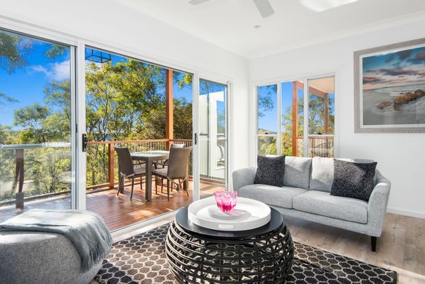 Bright contemporary living room with white walls, large floor-to-ceiling sliding glass doors, and a timber balcony overlooking leafy surroundings. The room contains a light grey sofa, a rounded black woven coffee table, a patterned dark rug, and a grey upholstered lounge chair in the foreground. A framed coastal landscape artwork hangs on the wall, while the adjoining outdoor dining balcony is visible through the glazing.