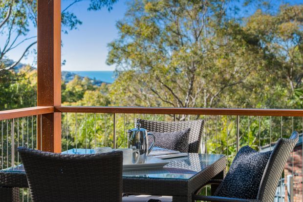 Outdoor dining terrace with dark woven wicker chairs and a glass-topped table, framed by warm timber posts and balustrading. The elevated setting overlooks leafy vegetation and a blue coastal or water vista under clear skies.