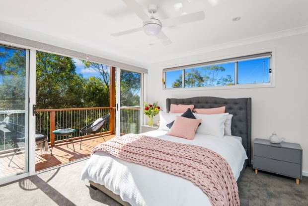Bright contemporary bedroom with a large upholstered bed, white bedding, blush pink cushions and knitted throw, dark grey bedside cabinet, grey carpet, ceiling fan, and extensive glazing opening onto a timber balcony surrounded by trees.