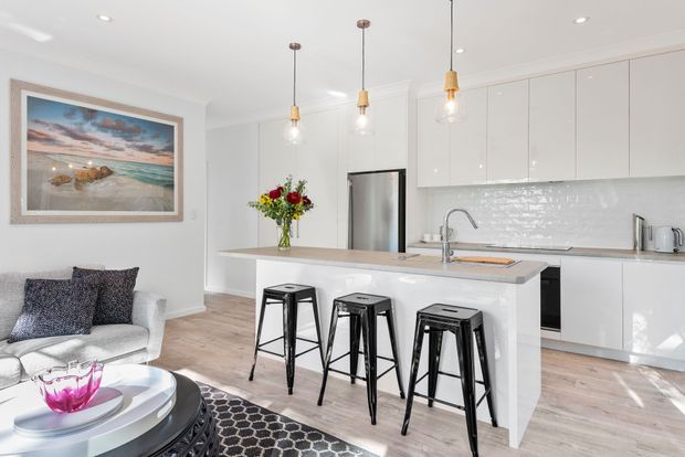 Bright, contemporary open-plan kitchen and living area with a large white island, pale timber-look flooring, glossy white cabinetry, white subway-tile splashback, stainless-steel appliances, and three black metal stools. A light upholstered armchair and patterned rug sit beside the kitchen, while pendant lights and a framed coastal artwork add decorative detail.
