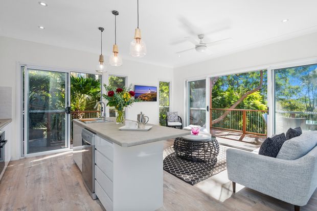 Bright open-plan kitchen and living area with a central white island, pendant lights, pale timber-look flooring, and large glazed doors opening onto a leafy deck. A light grey sofa, woven round coffee table, patterned rug, dining area, and indoor plants create a relaxed contemporary interior.