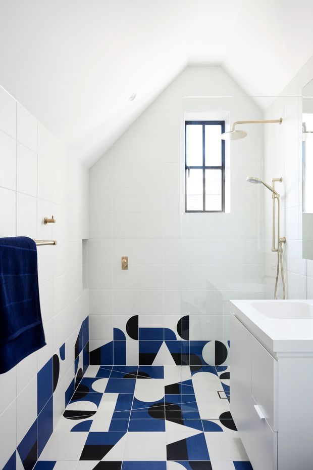Bright compact bathroom with a sloped white ceiling and white tiled walls. The floor and lower wall feature a bold geometric pattern in navy blue, black, and white. A freestanding-style white bathtub, glass shower enclosure, brass shower fittings, dark-framed window, white vanity, and deep blue towel create a contemporary graphic interior.