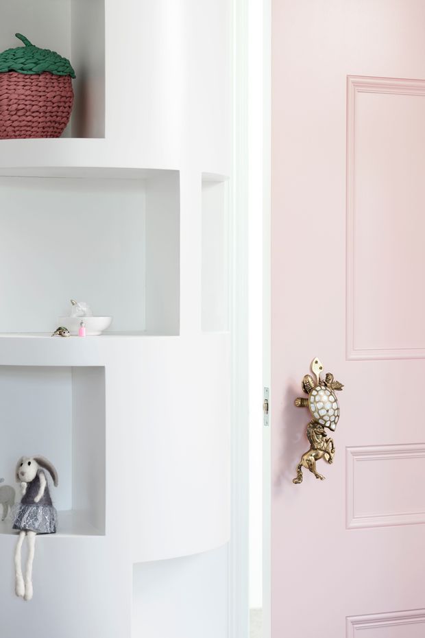 Bright, playful interior detail showing built-in white wall shelving with small decorative toys and objects, beside a pale pink paneled door fitted with an ornate gold turtle-shaped handle or door ornament. The space has a clean white-and-pastel palette and whimsical decorative styling.