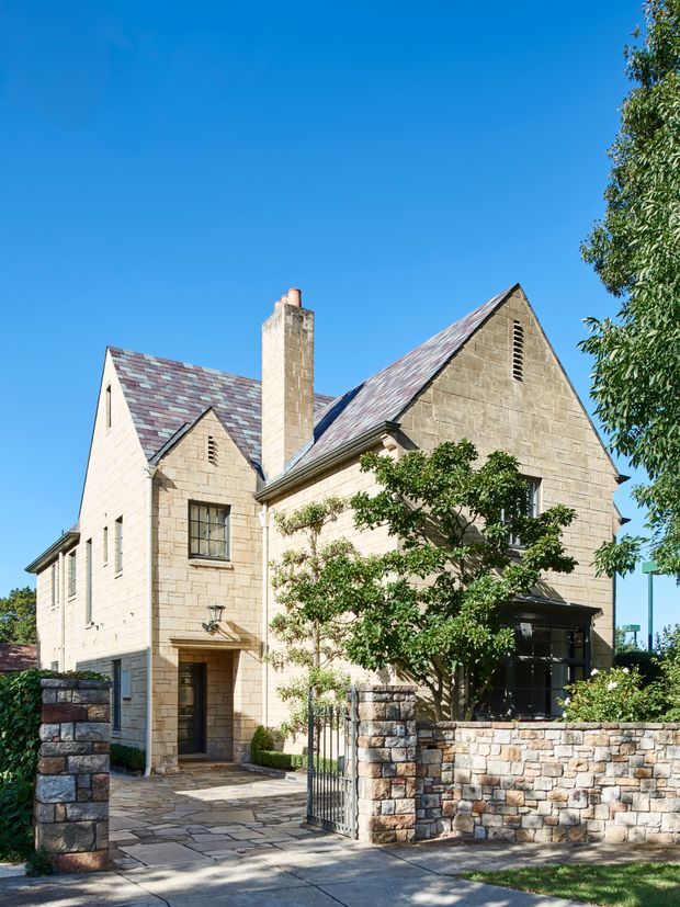 Exterior view of a substantial two-storey stone house with steep pitched gabled roofs, a prominent chimney, slate-like roof shingles, stone boundary walls, and a gated paved entry. The facade has traditional masonry detailing and small leaded-style windows, set among mature landscaping under a clear blue sky.