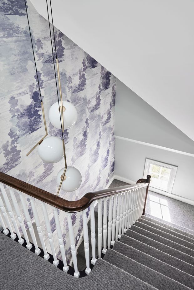 Interior view looking down a traditional stairway with white turned balusters, a dark polished timber handrail, grey carpeted stairs and landing, and a tall feature wall covered in lavender and white botanical-patterned wallpaper. Three spherical pendant lights hang through the stairwell void. A small white-framed window brings in daylight, while the sloping ceiling and angular wall geometry create an architectural focal point.