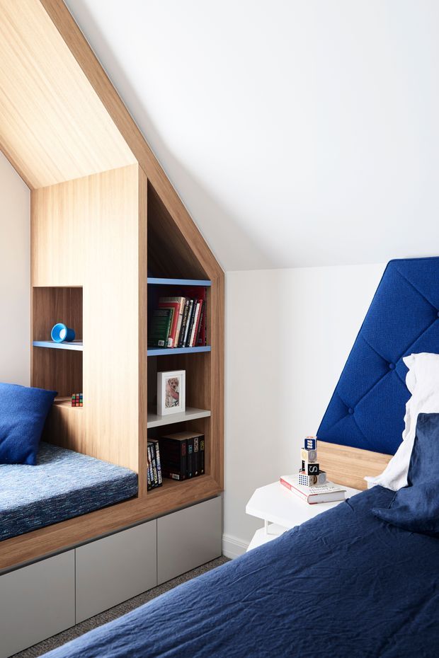 A compact, contemporary bedroom with built-in timber cabinetry and open shelving integrated beneath a sloped ceiling. The room features two blue-upholstered sleeping areas or beds, blue bedding, a white side table, books and small decor, with grey lower cabinetry and carpeted flooring. The palette is predominantly white, natural timber, grey, and deep blue.