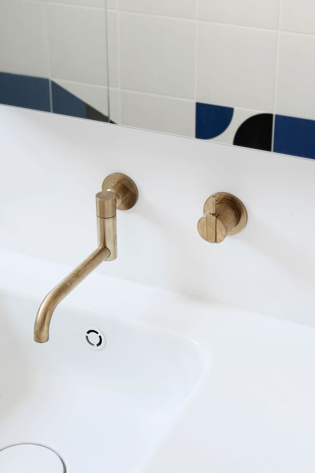 Close-up view of a minimalist bathroom basin with a white integrated or ceramic sink, wall-mounted brushed brass tap and separate control, and a mirror above. The upper wall is finished with white square tiles featuring a bold geometric accent band in navy blue and black. The composition is clean and contemporary with a restrained palette and distinctive graphic tiling.