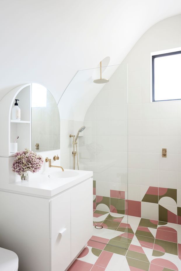 Bright contemporary bathroom with a white vanity and integrated basin, arched mirrored shelving, a frameless glass shower enclosure, pale wall tiles, and boldly patterned geometric floor and lower wall tiles in dusty pink, olive green, cream, and brown. Brass fixtures and a small high-set window add warmth and natural light.