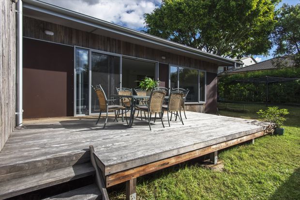 Exterior view of a low-profile contemporary house with a broad weathered timber deck, outdoor dining setting, dark brown wall panels, vertical timber cladding, sliding glass doors, and a gently sloping roof edge. The deck opens onto a grassed garden and is framed by mature trees and neighbouring residential structures.
