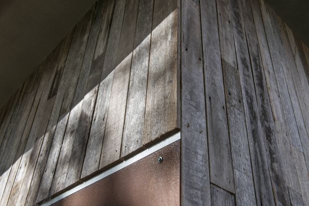 Close-up architectural detail of vertically arranged weathered timber wall cladding meeting at a corner, with a dark brown metal or painted panel at the lower edge. Strong angled sunlight creates high-contrast shadows across the textured wood surface.