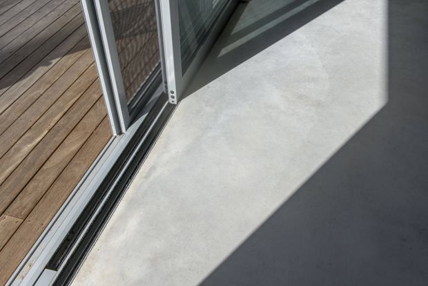 Close-up architectural detail of a large sliding glass door threshold opening onto a timber deck. The interior floor is a smooth pale grey concrete-like finish, with strong sunlight and sharp shadows cast across it. The door has slim silver aluminium framing and visible sliding tracks.
