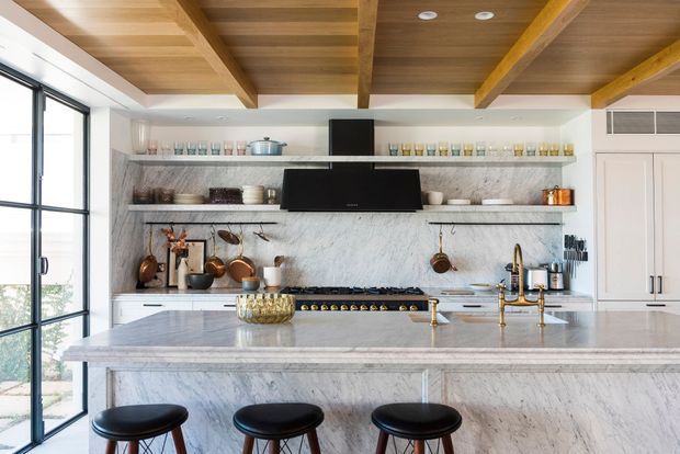A bright, refined kitchen with a large marble island and matching marble backsplash, white cabinetry, open display shelving, brass fixtures, and a black range hood. The room features a warm timber coffered ceiling with exposed beams, tall black-framed glazing, and three black counter stools.