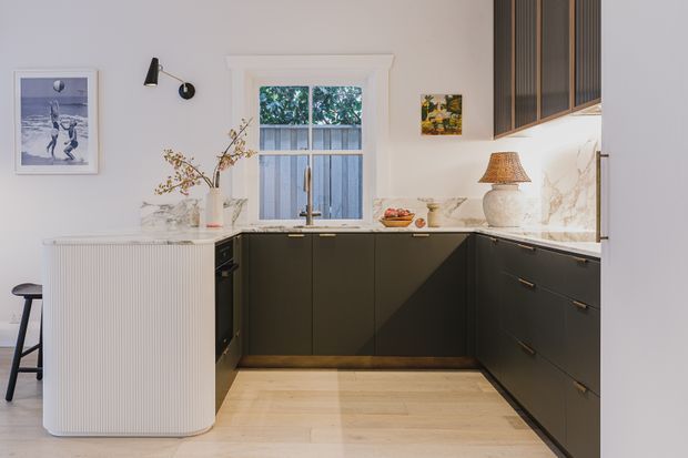 A compact, refined kitchen with a U-shaped layout, dark matte cabinetry, a pale stone benchtop and splashback, light timber flooring, and a curved fluted island or peninsula. White walls, a sash window, integrated under-cabinet lighting, and warm decorative accents create a bright contemporary interior.