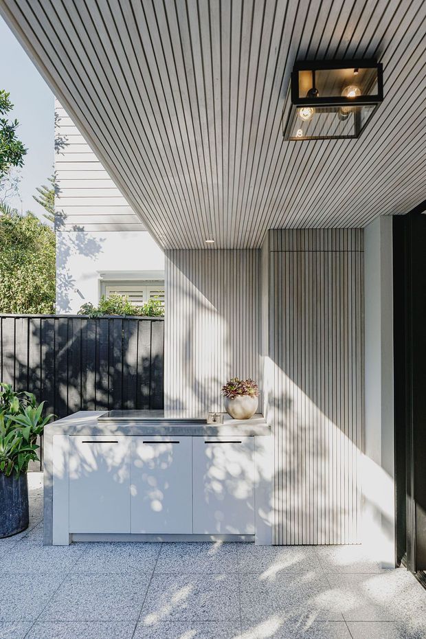 A bright contemporary covered outdoor entry or patio space with a full-height vertically ribbed wall and matching slatted ceiling. A white built-in storage cabinet with dark linear handles sits against the wall, topped with a stone-like surface and potted plant. Large-format grey terrazzo-like paving, black metal fencing, recessed lighting, and a black-framed glazed overhead light fixture complete the space. Lush planting and a neighbouring building are visible beyond the fence.
