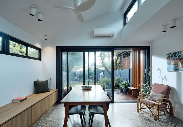 Bright contemporary dining area with a timber dining table and chairs, built-in timber sideboard, rattan lounge chair, ceiling fan, and large black-framed glazed doors opening to a landscaped outdoor space. White walls and ceiling, polished light terrazzo-like flooring, and abundant natural light create an airy connection between the interior and garden.