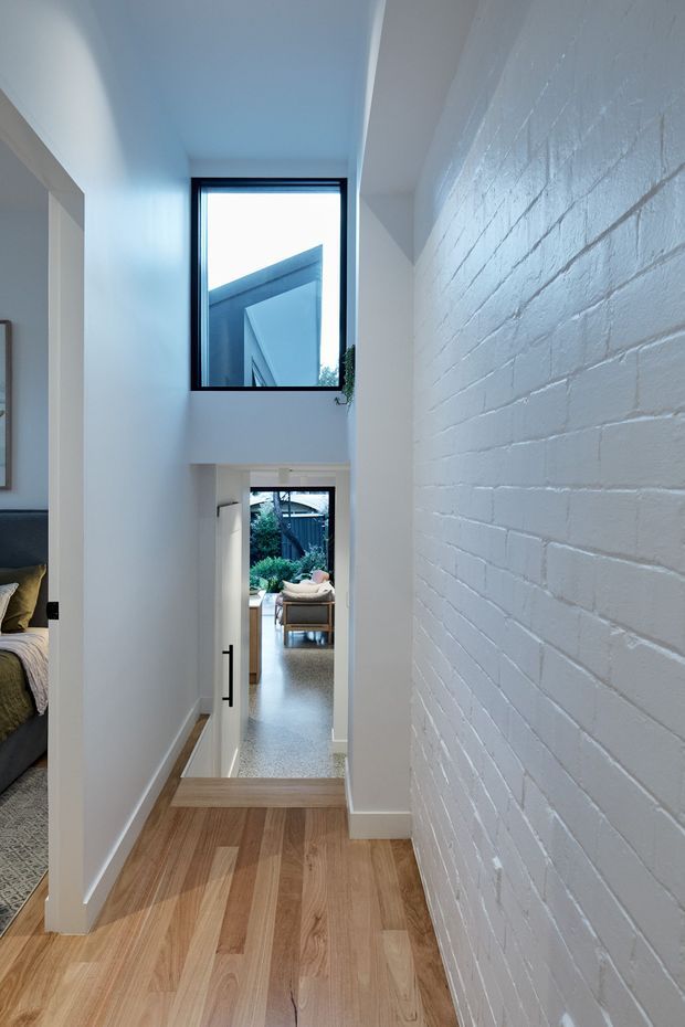 A bright, narrow interior hallway and stair landing with warm timber flooring, white-painted brickwork, white walls, and black-framed windows. A bedroom is partially visible through an opening on the left, while a glazed doorway at the end leads toward a furnished living area and greenery beyond.