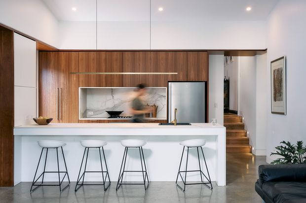 Contemporary open-plan kitchen dominated by a long white island with four slim black-framed stools. The kitchen features full-height dark timber cabinetry, a pale stone or marble-look backsplash, integrated stainless-steel appliances, polished concrete flooring, and a timber-lined stairway leading to an upper level. A partial black leather sofa and framed artwork are visible at the right edge.