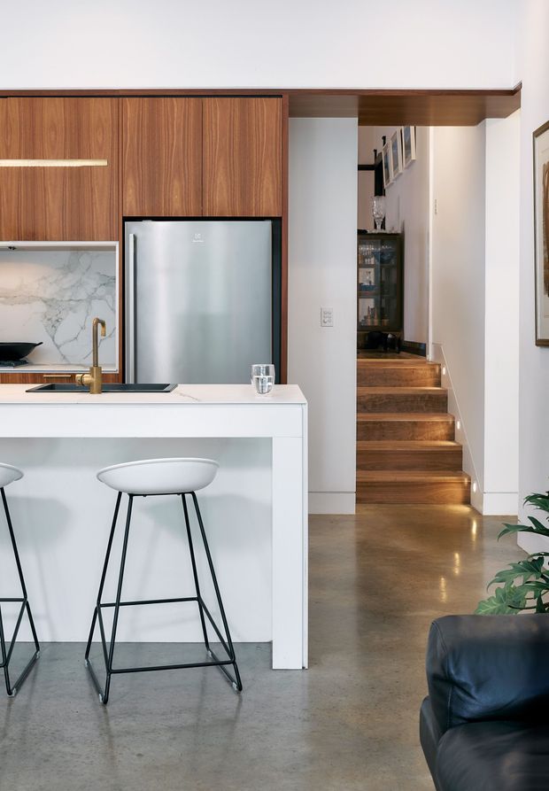 A modern open-plan kitchen viewed from the living area, featuring a white island with waterfall-style sides and a dark inset sink, walnut-toned full-height cabinetry, stainless-steel refrigerator, marble-look splashback, and brass tapware. Two minimalist bar stools sit at the island. A polished concrete floor extends toward a timber staircase leading to an upper level, with partial living-room seating and indoor planting visible at the right edge.