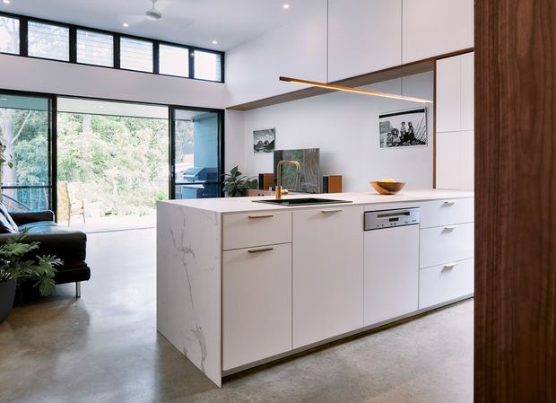 A bright, open-plan contemporary kitchen centred on a large white island with integrated sink, dishwasher and streamlined drawers. The island has a white marble-look surface and waterfall end panel, while full-height white cabinetry is paired with a warm timber feature panel at the right. Black-framed floor-to-ceiling glazing opens toward a leafy outdoor setting, and a slim linear pendant hangs above the island. A connected living area is visible in the background.