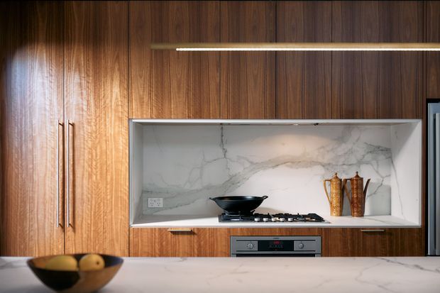 A close-up view of a refined contemporary kitchen featuring dark vertical-grain timber cabinetry, a white marble-look recessed cooking niche with integrated gas cooktop, built-in stainless-steel oven, and a matching pale stone island benchtop in the foreground. Warm linear lighting highlights the joinery and splashback.