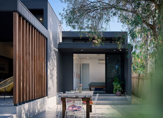 Modern residential courtyard viewed from outdoors, with dark charcoal cladding, vertical timber screening, and a glazed living or lounge space opening onto a paved patio. A timber dining table and wire chairs occupy the foreground, while trees and fencing frame the private outdoor setting.