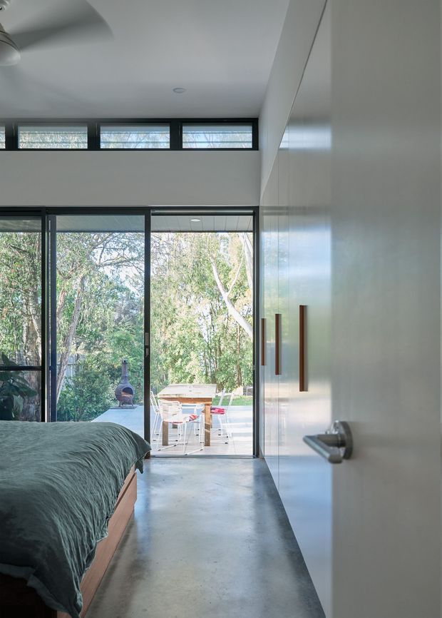A bright contemporary bedroom viewed toward full-height glazed doors opening onto a timber deck and leafy garden. The room features a low timber bed with dark green bedding, a polished concrete floor, white built-in cabinetry with long timber handles, black-framed glazing, and high clerestory windows. A compact outdoor dining setting and chiminea are visible beyond the glazing.