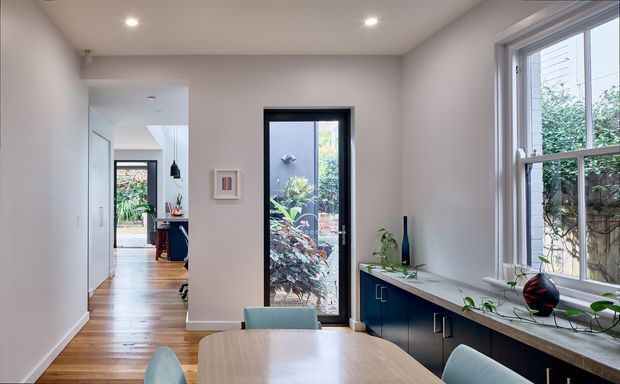 Bright contemporary dining area with a light timber dining table and upholstered chairs, opening to a kitchen beyond. The room features pale painted walls, recessed ceiling lights, a dark-framed glazed door, and a long built-in dark-blue cabinet beneath a large window with indoor plants. Timber flooring continues through the adjacent hallway toward an outdoor garden view.
