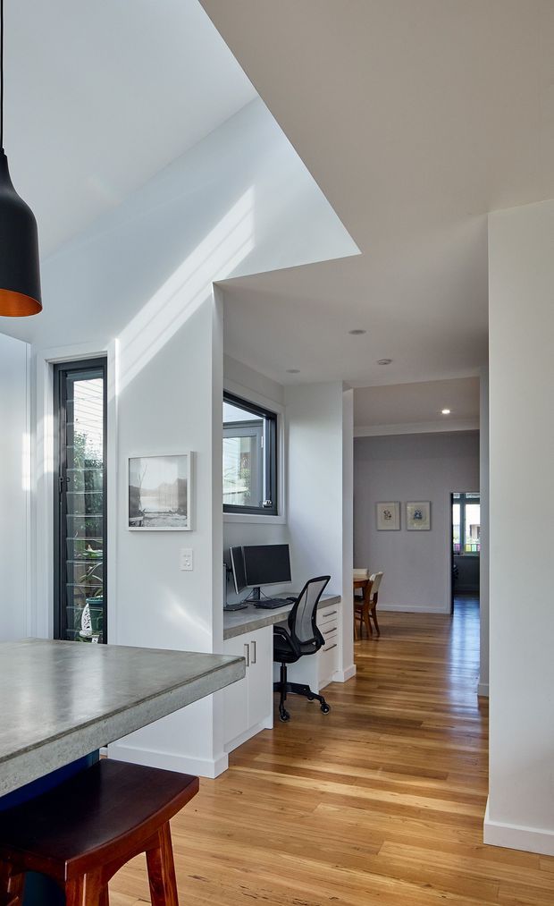 Bright contemporary open-plan interior showing a compact home office nook integrated beside a kitchen or breakfast counter. The space has white walls and ceiling, warm timber flooring, a grey concrete-look benchtop, white built-in drawers and cabinetry, dark-framed windows and doors, and a black pendant light. A hallway leads toward a dining area in the background. Strong daylight enters through the glazing and creates angular shadows across the walls.