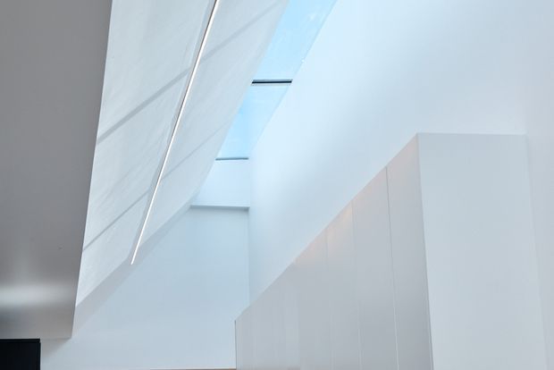 Minimalist interior view of a bright white stairway or landing, with a sculptural angled ceiling or wall plane, concealed linear detail, tall white partition walls, and narrow skylight openings bringing in cool natural light.
