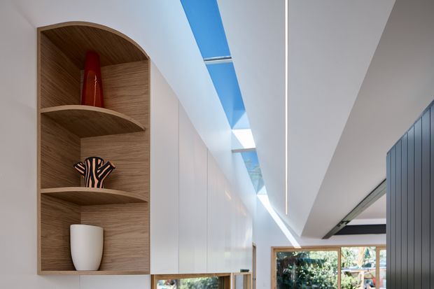 A contemporary interior corridor or open-plan living area with extensive white built-in cabinetry, a curved timber display niche with shelves, and a dramatic linear skylight creating a bright blue-lit slot in the ceiling. Glazing at the far end overlooks greenery, while a dark vertically ribbed wall or joinery element frames the right side. Minimal decorative objects add warmth to the otherwise clean, refined composition.