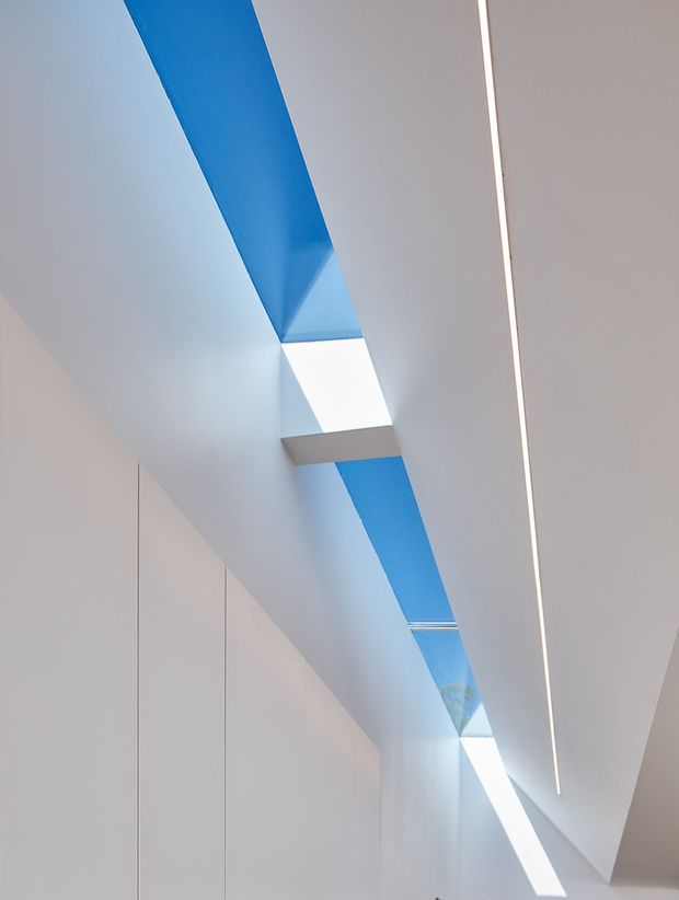 A minimalist interior view looking upward along a narrow passage or circulation space. Smooth white walls and ceiling are punctuated by a long linear skylight or roof opening revealing vivid blue sky, with a recessed white lightwell and a continuous slim linear light fitting. The composition is geometric, bright, and highly refined, with concealed doors or flush wall panels along the left side.