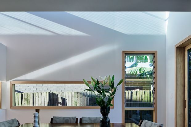 Bright contemporary interior corner with a high sloped ceiling, white walls, warm timber-framed windows and a glazed timber-framed doorway. Strong sunlight creates diagonal shadows across the walls and ceiling. A dining table with chairs and a vase of greenery occupies the foreground, while exterior fencing and planting are visible through the windows.