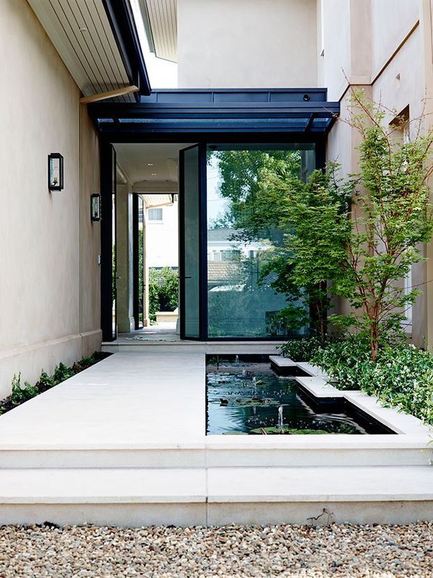 A refined residential entry courtyard viewed from outside, with broad pale stone steps and a smooth light-paved walkway leading through a tall glazed doorway. A dark metal-framed glass canopy and matching glazing create a strong contemporary architectural contrast against pale rendered walls. A narrow rectangular reflecting pond with small fountains runs alongside the path, bordered by dense planted greenery and a slender ornamental tree. The space is bright, calm, and highly composed, with a vertically enclosed courtyard character.