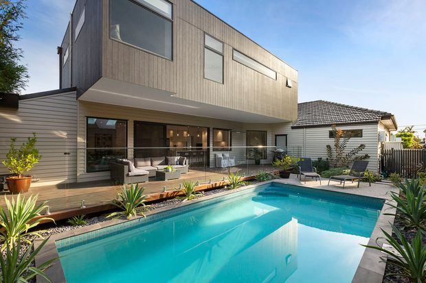 Exterior view of a contemporary two-storey home addition beside a swimming pool. The upper level is clad in vertical timber-look boards with large rectangular windows and projects over a glazed ground-floor living area. A timber deck, outdoor seating, established planting and the existing single-storey tiled-roof house are visible around the pool.