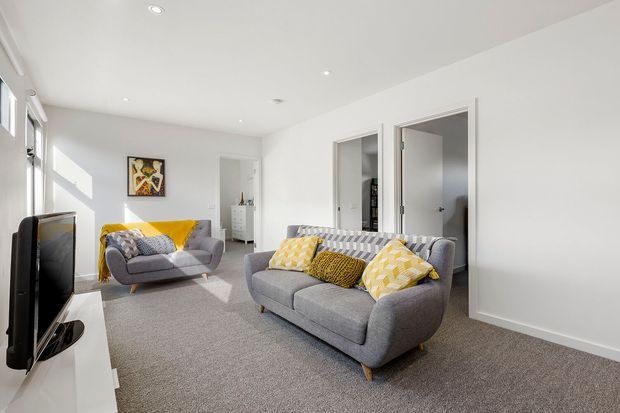 Bright, spacious living room with white walls and ceiling, grey carpet, and two grey upholstered sofas arranged around a television. Yellow and patterned cushions and a yellow throw add colour, while recessed ceiling lights, simple white door trims, and artwork create a clean contemporary interior. Sunlight enters through windows on the left.