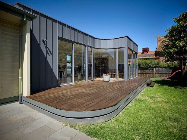 Exterior view of a contemporary single-storey house extension with a curved dark metal-clad facade, extensive floor-to-ceiling glazing, and a broad timber deck opening onto a landscaped backyard. The deck curves around the glazed living area, while an adjacent paved path and neighbouring residential structures are visible.