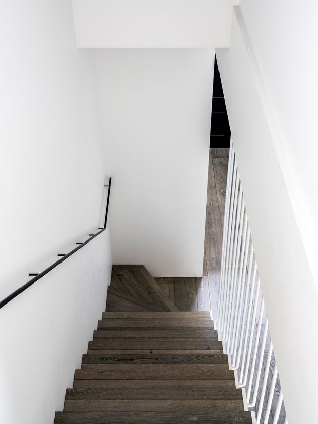 A bright, minimalist stairway viewed from above, with dark timber treads and landing, white walls, a slim black handrail, and a white vertical balustrade. The stairwell has a clean, contemporary architectural character and strong linear perspective.