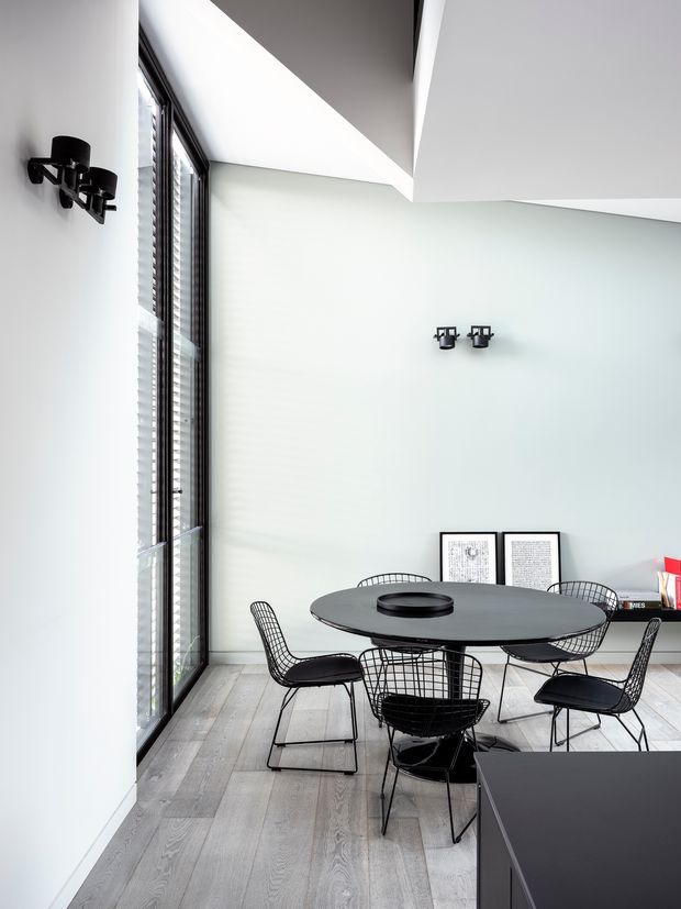 A bright, minimalist dining area with a round black dining table and wire-frame chairs positioned beside tall floor-to-ceiling glazing. White walls, a sculptural angular ceiling opening, pale timber flooring, black wall sconces, and sparse framed artwork create a crisp contemporary interior.