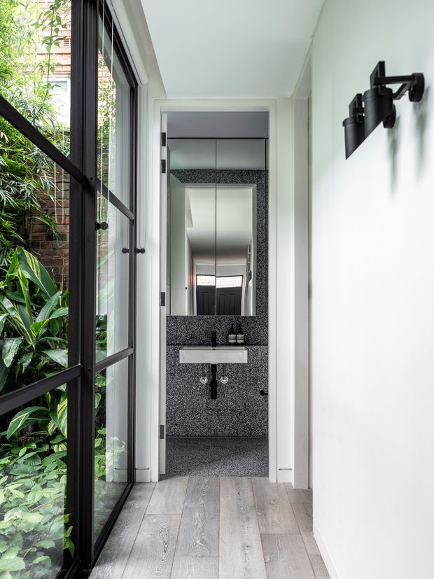 A bright, narrow hallway leading directly to a compact bathroom. The corridor has pale grey timber flooring, white walls and ceiling, a black-framed glazed door or window opening onto lush greenery, and minimalist black wall-mounted sconces. The bathroom features dark grey speckled stone or terrazzo wall and floor surfaces, a floating white rectangular basin, black fixtures, and a large mirrored cabinet.
