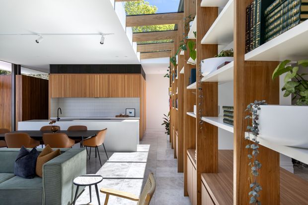 A bright, open-plan contemporary interior combining a kitchen, dining area and living space. The kitchen features a white island and cabinetry, a pale square-tile splashback, dark timber vertical wall panelling and a black tap. A timber-framed overhead skylight or clerestory structure brings in natural light. Floor-to-ceiling timber shelving and joinery line the right side, displaying books and plants. The foreground includes a muted green sofa, brown leather dining chairs and a low round side table. The palette is warm timber, white, grey and muted green, with a strong indoor-outdoor feel from the overhead glazing.