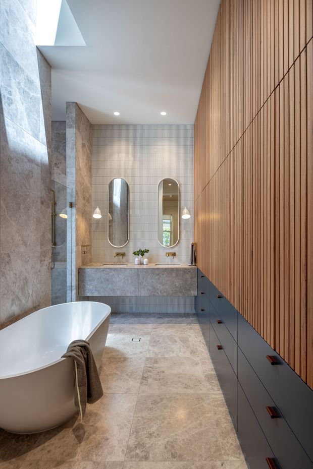 A refined contemporary bathroom with a freestanding white bathtub, double vanity, arched mirrors, wall-mounted pendant lights, and extensive built-in storage. Large stone-look slabs cover the left wall and floor, while a vertical timber slat feature wall adds warmth. The vanity wall is finished with small-format light tiles, and a skylight brings in natural light.
