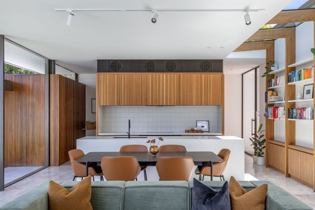 A bright, open-plan contemporary kitchen and living area. The kitchen features a long white island with an integrated sink, white tiled splashback, timber slatted overhead cabinetry, and warm timber wall panelling. A dining table with brown upholstered chairs sits in front of the island, while a large sofa occupies the foreground. Floor-to-ceiling glazing, timber screening, exposed timber ceiling beams, open shelving, and built-in cabinetry create a warm, refined interior.
