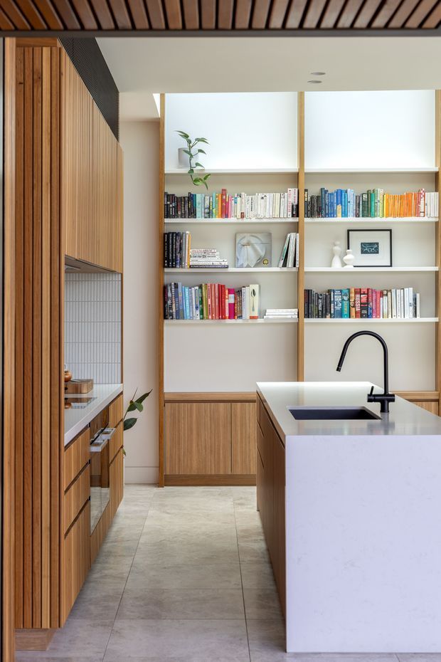 Contemporary galley-style kitchen with extensive vertical timber cabinetry, a large pale stone island with an integrated dark sink and curved black tap, and built-in open shelving filled with books and a few decorative objects. The kitchen has a warm timber slatted ceiling detail, light walls, pale large-format tiled flooring, and a clean, refined composition with abundant storage.