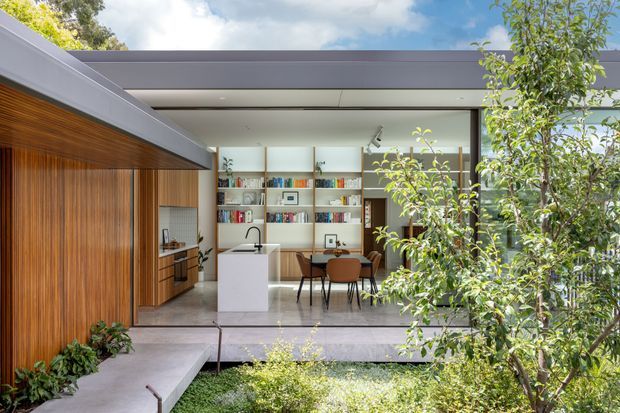 View from a landscaped courtyard toward a contemporary single-storey home. The open-plan interior features a white kitchen island, timber-lined cabinetry and wall, a dining table with brown chairs, and extensive built-in shelving. Broad roof overhangs, dark horizontal fascia elements, large openings, and a sheltered outdoor threshold create a strong indoor-outdoor connection.