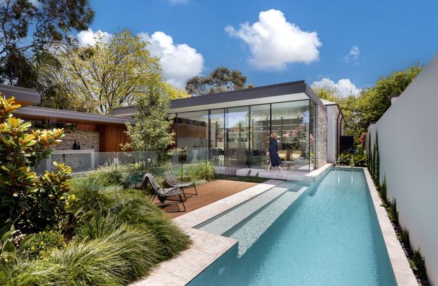 A contemporary residential courtyard viewed from the outdoor garden area, featuring a long narrow swimming pool beside a timber deck and landscaped planting. A glazed pavilion or living space with a sharply angled roof and dark metal framing opens directly onto the pool, while a low brick-clad building and pale boundary walls enclose the setting. The scene is sunlit with a clean, indoor-outdoor connection and strong geometric lines.