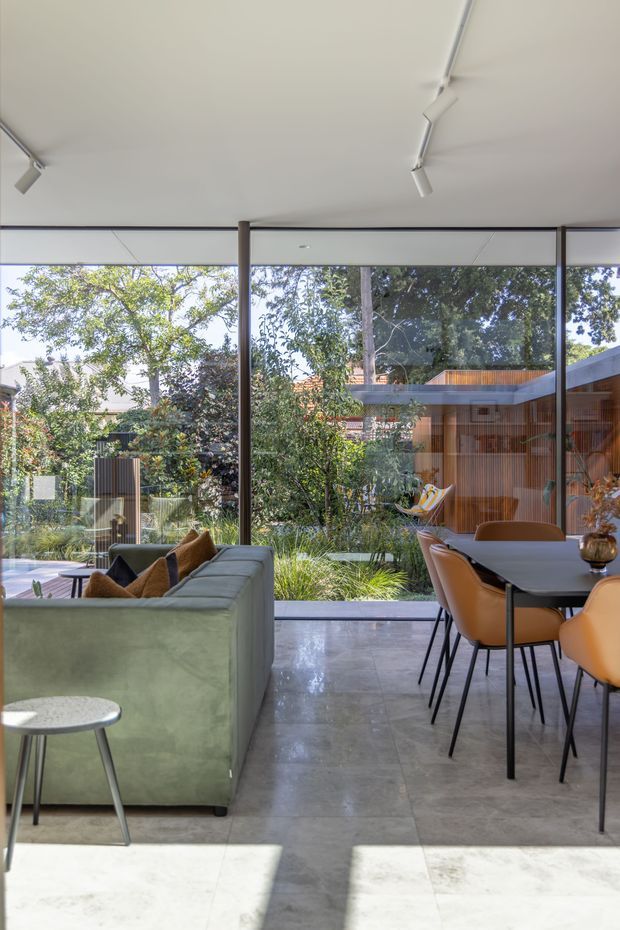 A bright, modern open-plan living and dining space with full-height glazing overlooking a lush planted courtyard. The room features a pale green sofa, dark dining table with tan leather chairs, polished grey stone or concrete-look flooring, a white ceiling with track lighting, and slim dark window frames. Timber-clad garden structures are visible beyond the glazing.