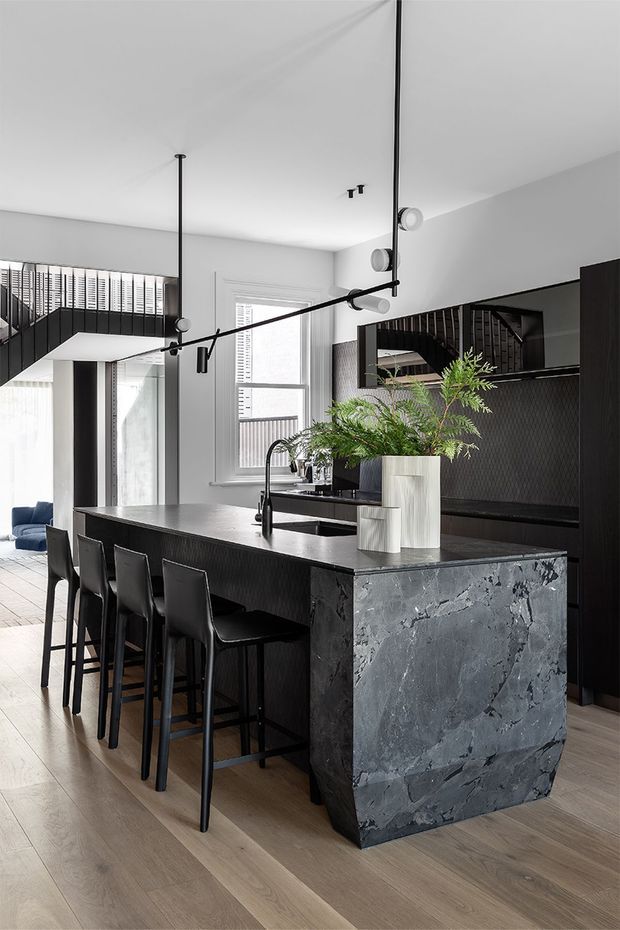 A contemporary open-plan kitchen dominated by a large dark island with seating for four. The island has a dramatic grey natural-stone waterfall end and dark textured cabinetry, complemented by a black tiled or patterned splashback, black tapware, and integrated appliances. Pale timber flooring, white walls and ceiling, large windows with blinds, a staircase, and black linear pendant lighting create a high-contrast monochromatic interior. A living area is partially visible beyond the kitchen.